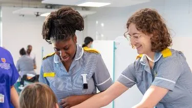 Two female healthcare students in grey uniforms assist a seated person in hospital